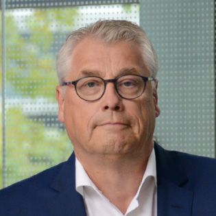 Mann in dunklem Anzug lehnt an einem Gel&auml;nder in einem modernen Geb&auml;ude mit gro&szlig;en Fenstern