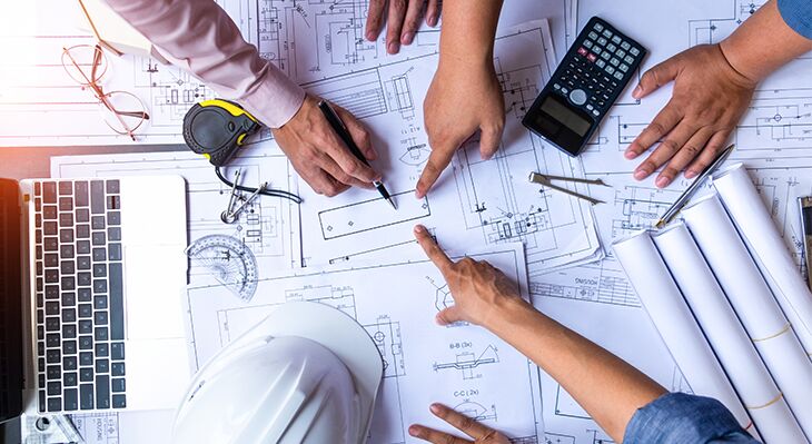 H&auml;nde, die auf Baupl&auml;ne zeigen. Helm, Laptop und weitere kleine Gegenst&auml;nde liegen rund um die Pl&auml;ne verteilt. 