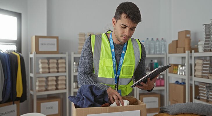 Junger Mann in einer Warnweste, der ein Kiste packt; Tablet in der Hand. 