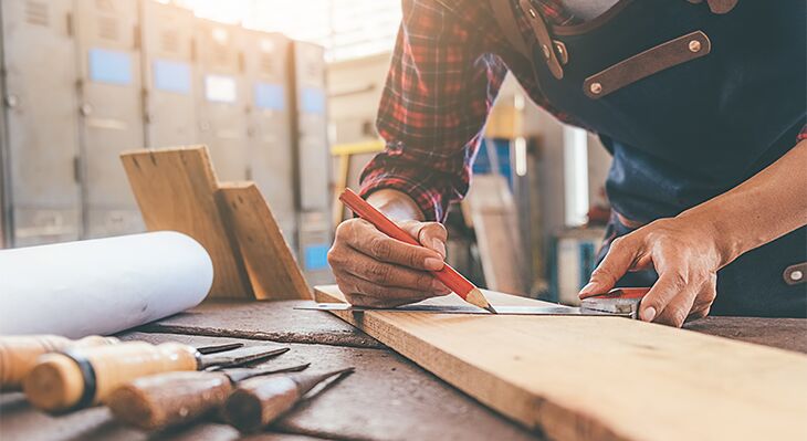 Mann in blauer Sch&uuml;rze, der ein Brett ausmisst.  