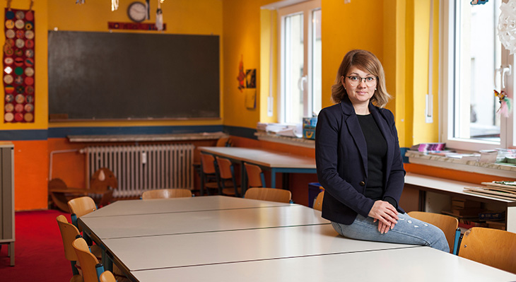 Eine Lehrerin sitzt in einem leeren Klassenraum auf einem Tisch, hinter ihr sieht man die Tafel.