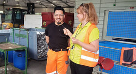 Two people in a warehouse; one is wearing orange work trousers and a black T-shirt, the other is wearing a yellow waistcoat and is holding a microphone.