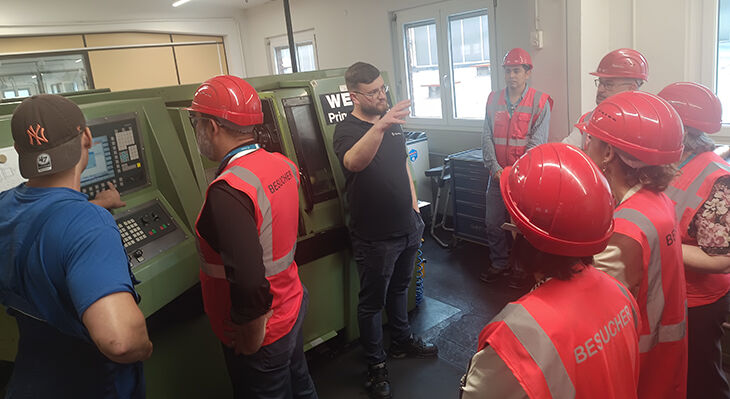 A group of visitors wearing red waistcoats and helmets stand in front of a young man in a black T-shirt, who is standing in front of a machine and explaining something to the group.