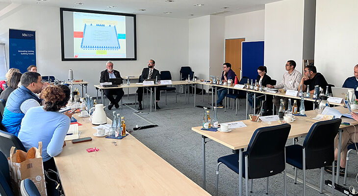Conference room with around 13 people; at the front, two men are seated in front of a PowerPoint presentation