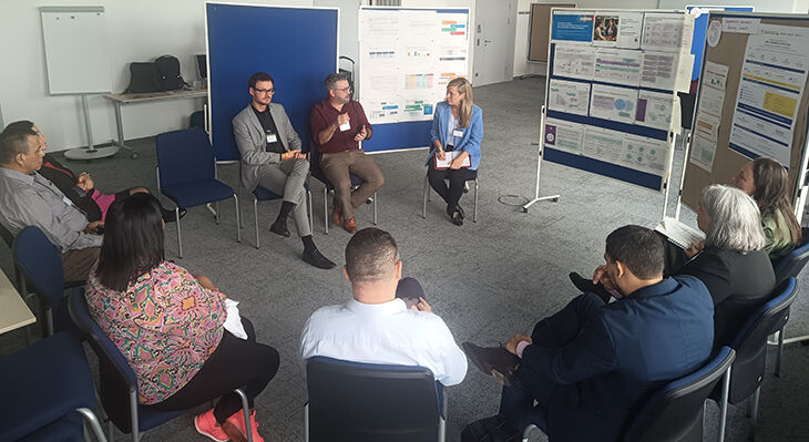 A circle of ten people seated; in the background are noticeboards displaying numerous diagrams and other printed slides