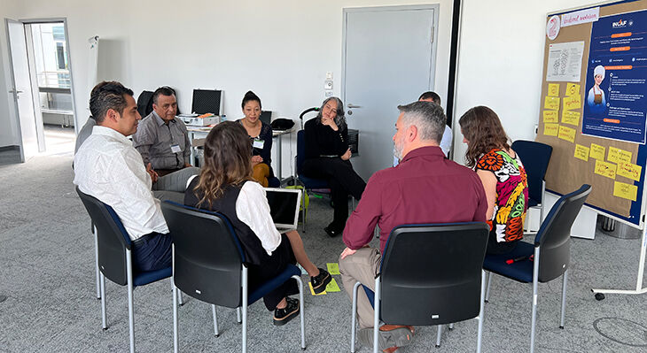 A circle of ten people seated; in the background are a poster and several labelled Metaplan cards