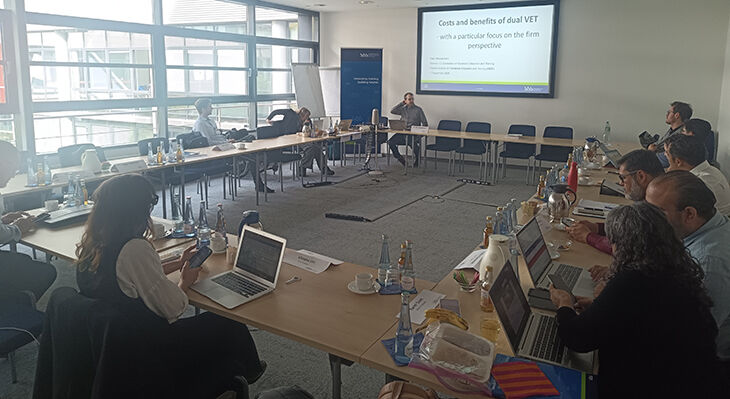 Conference room with around 15 people; at the front, a man is seated in front of a presentation entitled ‘Costs and benefits of dual VET’.