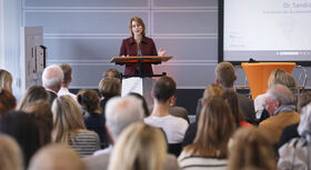 Frau in Blazer spricht an einem Rednerpult vor sitzendem Publikum in einem Konferenzraum.