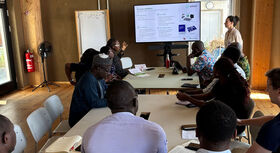 Several people are sitting at a long table in a room, looking at a presentation on a screen