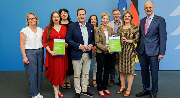 Gruppe von neun Personen steht vor blauem Hintergrund mit EU- und Deutschlandflagge, zwei Frauen halten gr&uuml;ne Ordner