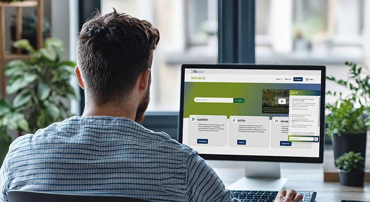 Mann mit Bart und blauem Hemd sitzt vor Laptop mit ge&ouml;ffneter Webseite in modernem B&uuml;ro mit Pflanzen im Hintergrund