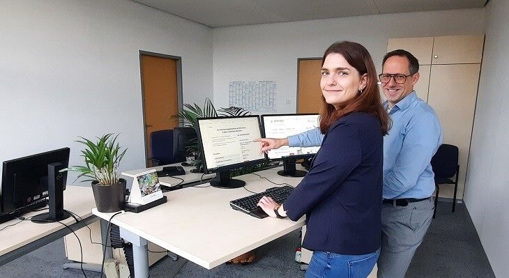 Zwei Personen stehen an einem Stehpult mit zwei Bildschirmen, eine Person zeigt auf einen Bildschirm.