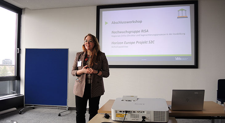 Eine Frau steht in einem Seminarraum vor einer Pr&auml;sentationsfolie und spricht. Auf der Leinwand steht ‚Abschlussworkshop'