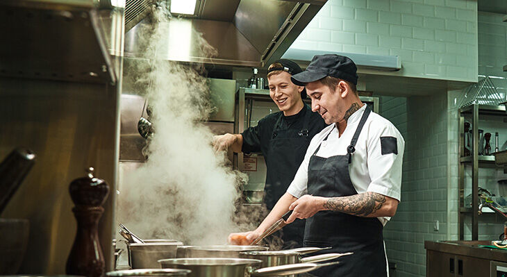 Zwei K&ouml;che in Kochkleidung arbeiten in einer professionellen K&uuml;che mit dampfenden T&ouml;pfen auf dem Herd