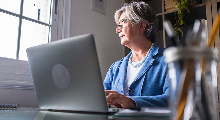 Ältere Frau mit grauen Haaren sitzt an einem Schreibtisch und arbeitet an einem Laptop vor einem Fenster.