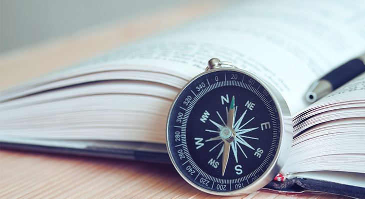 Runder Kompass mit schwarzem Zifferblatt und silbernem Rand liegt auf offenem Buch mit sichtbarem Text, daneben ein Stift.