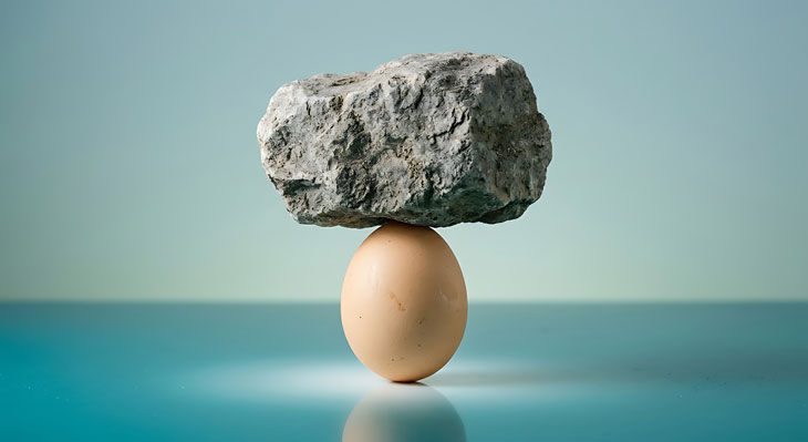 Ein gro&szlig;er Stein balanciert auf einem Ei, das auf einer glatten Oberfl&auml;che steht