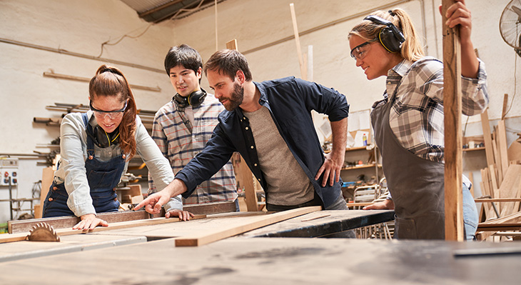 Drei Personen in Arbeitskleidung stehen um einen Holztisch in einer Werkstatt, eine Person zeigt auf ein Holzbrett.