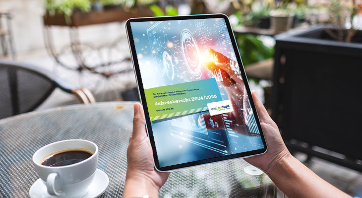 Person h&auml;lt Tablet mit digitaler Anzeige und Text 'Jahresbericht 2024/2025' vor einem Tisch mit Kaffeetasse