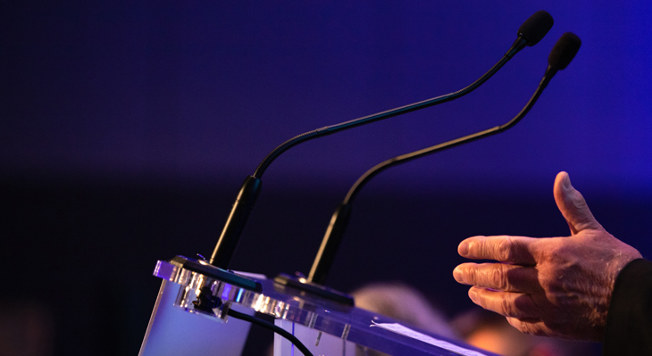 Zwei schwarze Mikrofone auf einem Podium. Der Hintergrund ist dunkel und lila. Im Vordergrund eine männliche Hand.