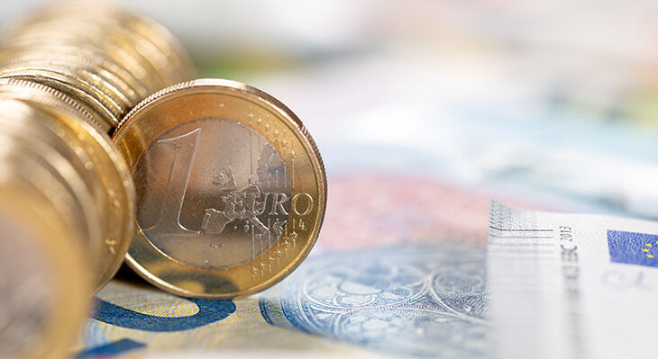 Ein Stapel Euromünzen liegt auf der Seite, eine der Münzen ist so aus dem Stapel gerollt, dass man die Vorderseite gut sehen kann. Die Münzen liegen auf Euroscheinen, die man nur verschwommen erkennt.