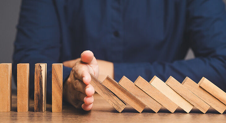 Hand stoppt fallende Dominosteine auf einem Holztisch vor einer Person in blauem Hemd
