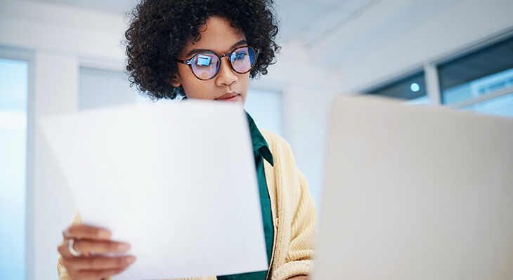 Frau mit lockigem Haar und Brille liest konzentriert ein wei&szlig;es Blatt Papier in einem modernen B&uuml;ro