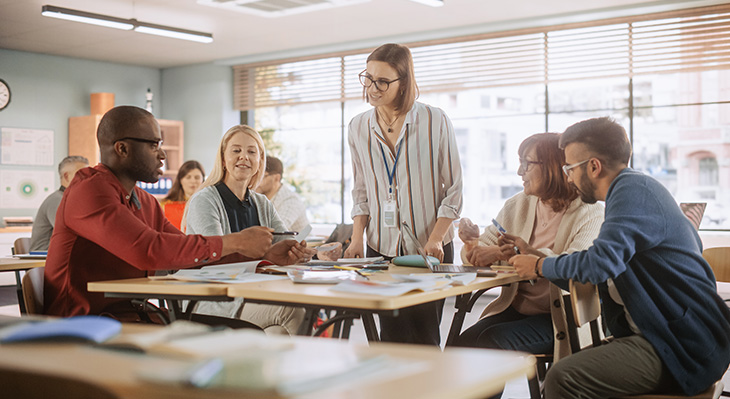 Unterricht an einer Volkshochschule: Erwachsene Teilnehmende lernen gemeinsam in zwei Teams und nehmen an einem sprachlichen Lernspiel teil. Die Lehrkraft ber&auml;t, beantwortet Fragen und motiviert die Gruppe.