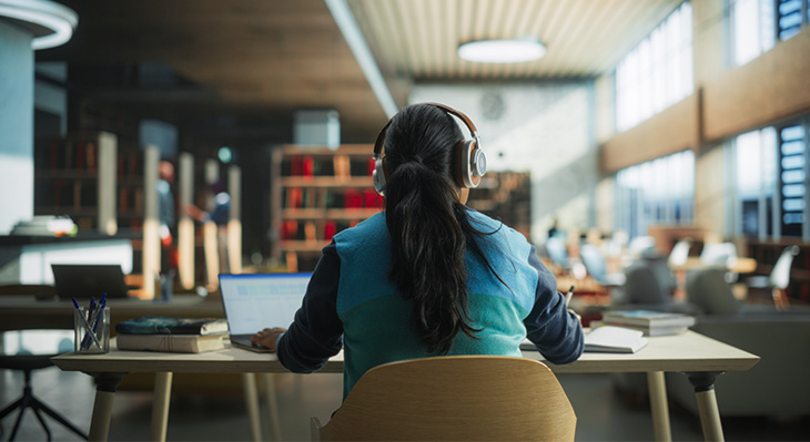 Frau mit Zopf und dunkelbraunen Haaren sitzt mit dem Rücken zur Kamera. Sie trägt Kopfhörer und sitzt an einem Tisch in einer Bibliothek. Auf dem Tisch steht ein Laptop und sie schreibt etwas in ihren Notizblock. 