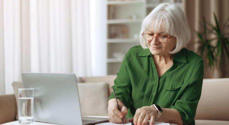 &Auml;ltere Frau mit Brille und gr&uuml;nem Hemd sitzt am Laptop und schreibt auf Papier