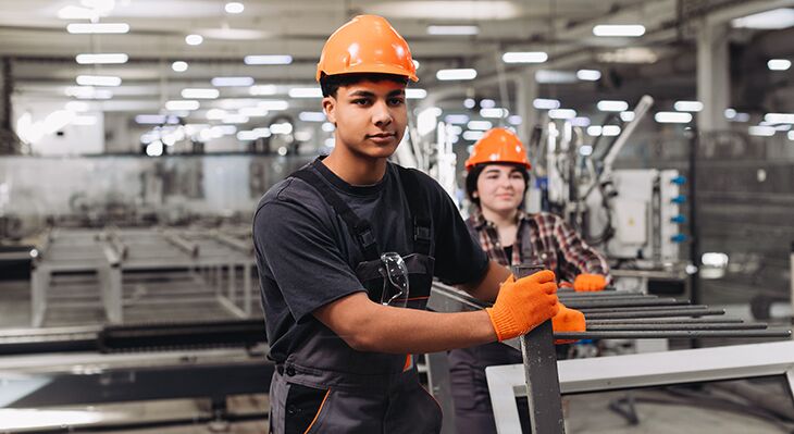 Zwei Auszubildende in einer Industriehalle, die an einem Metallrahmen arbeiten.