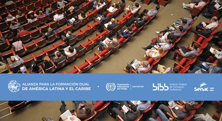 Konferenzsaal von oben fotografiert, Sitzreihen, davon einige Sitze unbesetzt. Darüber Schriftzug mit Logos, unter anderem BIBB und Allianz für duale Berufsbildung Lateinamerika.