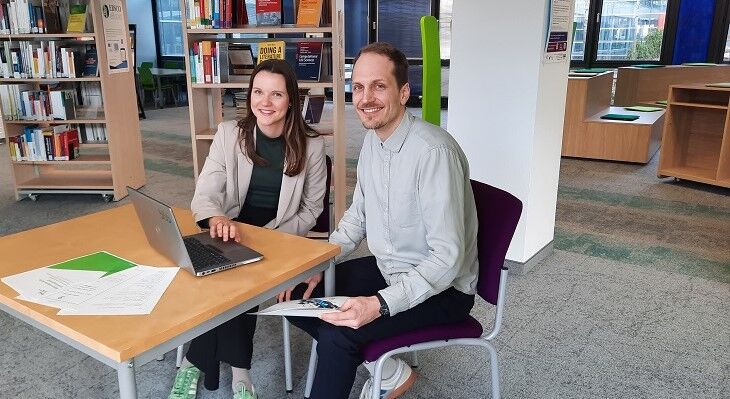 Nora Rohr (links) und Moritz Scholz (rechts) sitzen an einem Tisch mit einem Laptop und l&auml;cheln in die Kamera