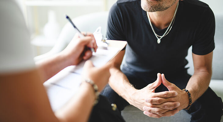 Ein Mann sitzt vor einer Person, die auf einem Fragebogen schreibt.
