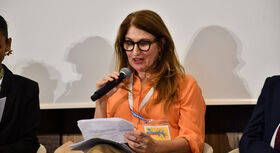 Frau mit langem Haar, orangefarbener Bluse und Namensschild h&auml;lt Mikrofon und Papiere in der Hand, sitzt auf einem Podium.