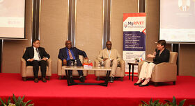 Vier Personen sitzen in Sesseln auf einer Bühne mit rotem Teppich beim Policy-Dialog.