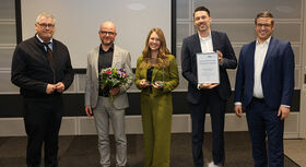 F&uuml;nf Personen stehen nebeneinander vor einer grauen Wand, eine h&auml;lt einen Blumenstrau&szlig;, eine eine Urkunde, eine weitere eine kleine Auszeichnung.