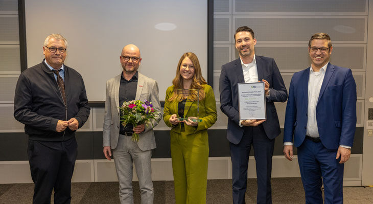 F&uuml;nf Personen stehen nebeneinander vor einer grauen Wand, eine h&auml;lt einen Blumenstrau&szlig;, eine eine Urkunde, eine weitere eine kleine Auszeichnung. 