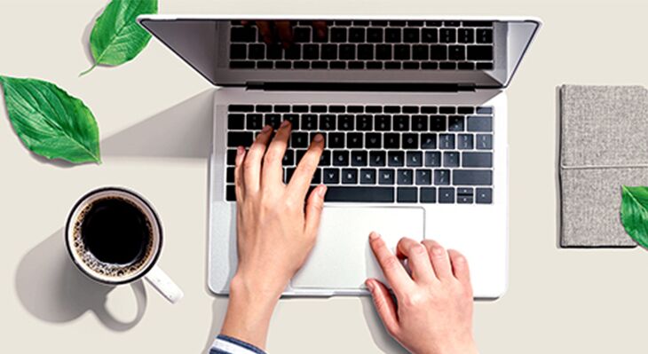 Draufsicht auf einen Laptop mit tippenden H&auml;nden, daneben eine Tasse schwarzen Kaffees, gr&uuml;ne Pflanzenbl&auml;tter und ein graues Notizbuch auf hellem Hintergrund.