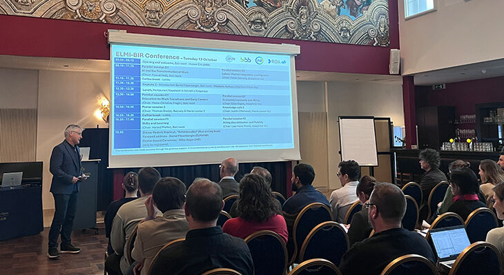 Mann mit Brille steht vor sitzendem Publikum neben einer Leinwand mit Konferenzprogramm in einem Saal mit gro&szlig;em Wandgem&auml;lde und hohen Fenstern.