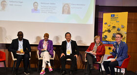 F&uuml;nf Personen sitzen auf einer B&uuml;hne in Sesseln, eine Person h&auml;lt ein Mikrofon, im Hintergrund ein Bildschirm mit Text und ein gelbes UNESCO-Banner.