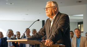 Mann in Anzug steht an Rednerpult mit Mikrofon vor einer Gruppe von Menschen in einem Innenraum