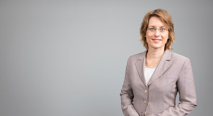 Frau mit kurzen Haaren und Brille im hellen Blazer steht vor neutralem Hintergrund und l&auml;chelt in die Kamera.