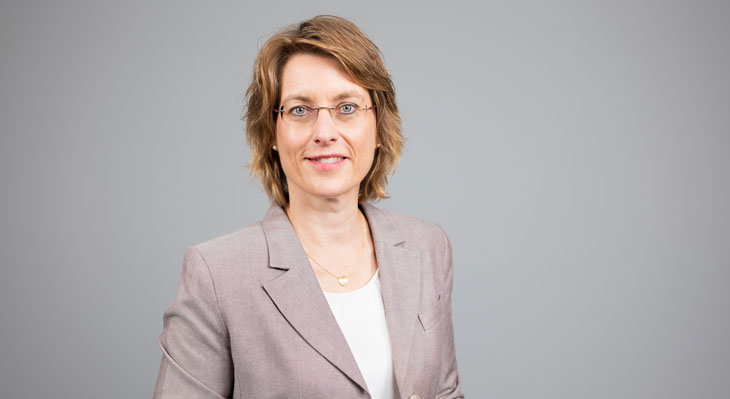 Frau mit kurzem, welligem Haar und Brille in einem hellen Blazer vor grauem Hintergrund