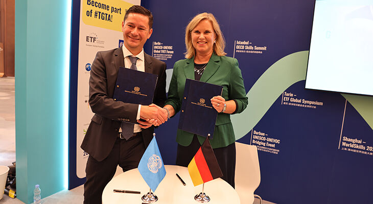 Ein Mann im dunklen Anzug und eine Frau im gr&uuml;nen Blazer geben sich bei der WorldSkills Conference Shanghai 2026 die Hand und pr&auml;sentieren unterzeichnete Vereinbarungen.