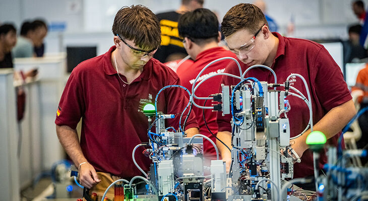 Zwei deutsche Teilnehmer der WorldSkills Shanghai 2026 arbeiten konzentriert an einer automatisierten Industrieanlage mit zahlreichen Kabeln, Sensoren und Steuerungselementen.
