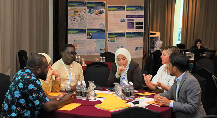 Fünf Personen sitzen an einem runden Tisch mit roter Tischdecke und haben Unterlagen und Wasserflaschen vor sich.