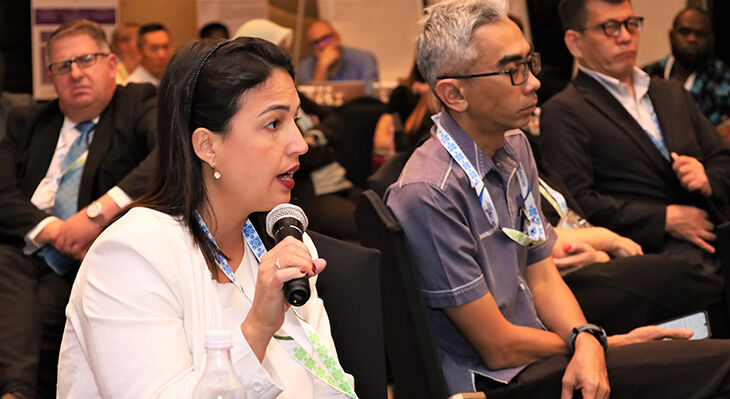 Eine Frau in weißer Bluse sitzt im Publikum und hält ein Mikrofon und ein Smartphone in den Händen, umgeben von weiteren Personen.