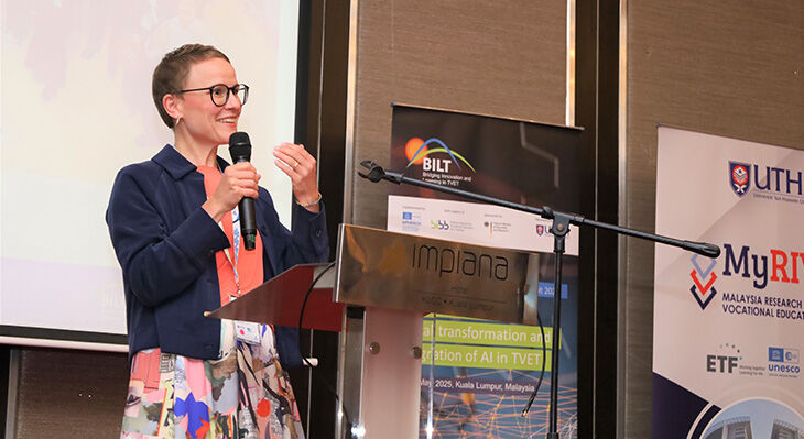 Eine Frau mit Mikrofon steht am Rednerpult , trägt eine dunkelblaue Jacke und spricht bei Veranstaltung mit Bannern im Hintergrund.