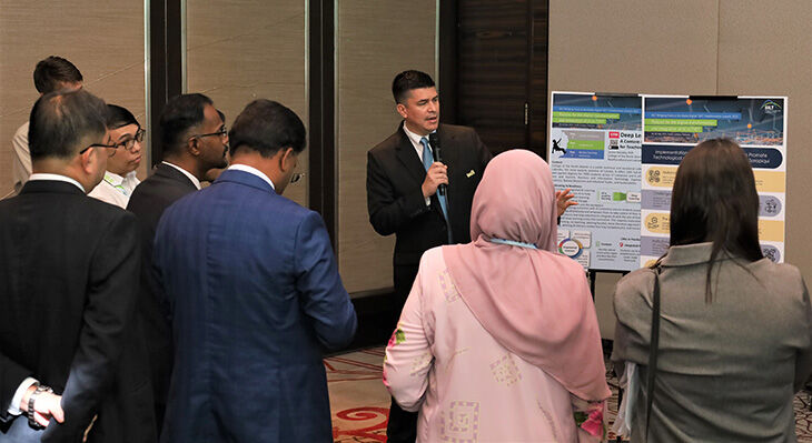 Eine Person im Anzug präsentiert ein Poster vor einer Gruppe von sieben Zuhörern in einem  Konferenzraum.